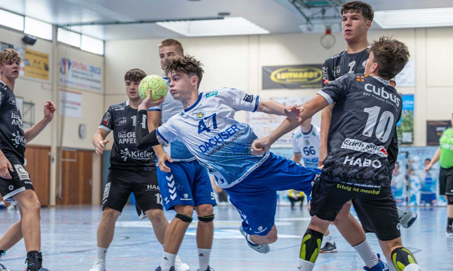Handballspieler im weißen Trikot springt zum Wurf, zwei Gegner blocken.