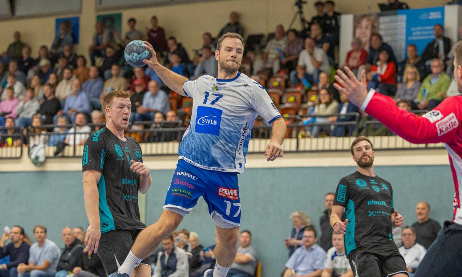 Spieler im weißen Trikot springt zum Wurf, zwei Gegner blocken.