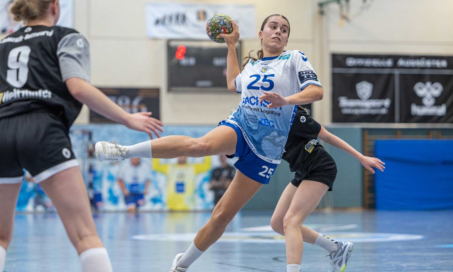 Spielerin im weißen Trikot springt zum Wurf, zwei Gegnerinnen blocken.